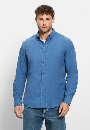 Mann mit lockigem Haar, der ein blaues Hemd mit Knöpfen und blaue Jeans trägt und vor einem einfarbigen hellen Hintergrund steht.