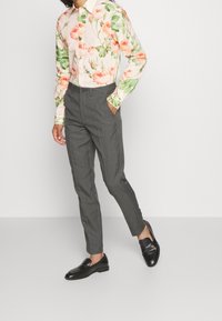 Persona vestida con pantalones grises a medida, mocasines negros y una camisa de manga larga con un patrón de rosas rosas y hojas verdes, con una mano en el bolsillo.