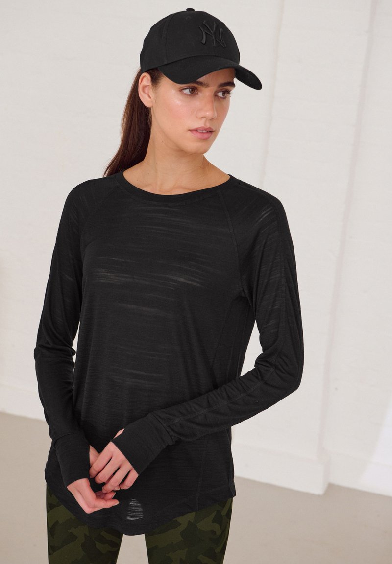 Schwarzes Langarm-Performance-Shirt mit Rundhalsausschnitt und Daumenlöchern, kombiniert mit Leggings im Camouflage-Muster und einer schwarzen Baseballkappe.