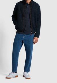 Fleecejacke in Dunkelblau mit Reißverschluss und schwarzem Besatz, kombiniert mit mittelblau gewaschenen, konisch zulaufenden Jeans und weißen Turnschuhen.