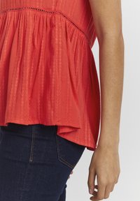 Top peplum rojo con rayas verticales y un acabado texturizado, con un dobladillo acampanado. Combinado con jeans azul oscuro.