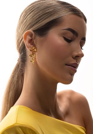 Ženska s svetlo rjavimi lasmi, zvezanimi nazaj, ki nosi zlate cvetlične viseče uhane in rumeno majico brez ramen, z zaprtimi očmi, obrnjena v desno.