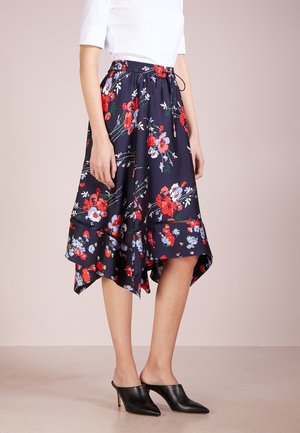 Vrouw draagt een marineblauwe midi-rok met rood-witte bloemen, gecombineerd met zwarte hakmuiltjes en een wit top met driekwart mouwen.