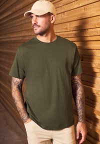 Camiseta de manga corta verde oliva, con escote redondo, de material de algodón suave, combinada con pantalones beige, de pie frente a un fondo de madera.