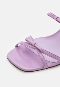 Sandale en cuir verni lilas avec bout carré, brides croisées et petit nœud près de la zone des orteils.