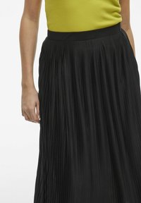 Jupe plissée noire en tissu texturé. La taille haute et les plis verticaux créent un design fluide. Portez-la avec un haut jaune.