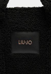 Materiale in faux fur nero con un'etichetta in pelle nera che presenta il marchio "LIU·JO" in oro, in rilievo. Aspetto morbido e texture piacevole.