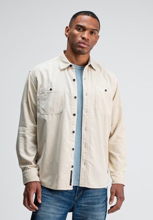 Beige overhem met knopen en twee borstzakken, zwarte knopen en een ontspannen pasvorm, gedragen over een lichtblauw T-shirt. Ggedragen met blauwe spijkerbroeken.