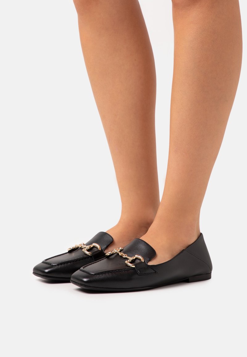 Femme portant des mocassins en cuir noir avec un détail chaîne dorée, debout sur un fond blanc uni.