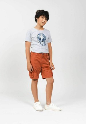 Grijze T-shirt met een blauwe doodskop graphic; gecombineerd met oranje cargoshorts met zijzakken; witte sneakers maken de outfit af.