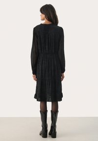 Zwarte jurk met lange mouwen en een gelaagd ontwerp, voorzien van een ruchesnek en een elastische taille, gecombineerd met zwarte laarzen tot de knie.