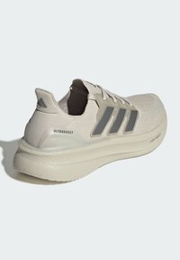 Zapatilla de running Adidas Ultraboost beige con parte superior de tejido, tres franjas grises en el lateral y suela gruesa acolchada etiquetada como Lightboost.