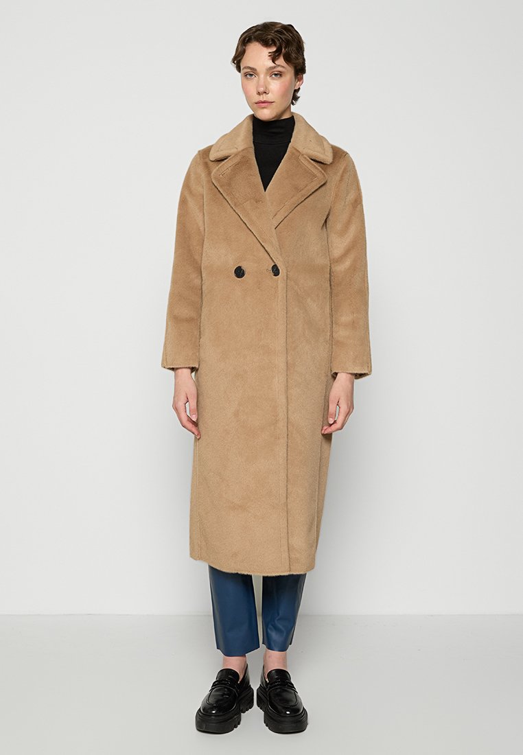 Kobieta ubrana w długi beżowy płaszcz dwurzędowy, czarny golf, niebieskie spodnie typu crops i czarne buty na platformie, stojąca przy białej ścianie.