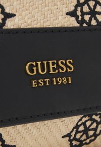 Bolso de textil negro y paja con el logo dorado "GUESS EST 1981". Presenta intrincados patrones negros en la paja y acentos de cuero.