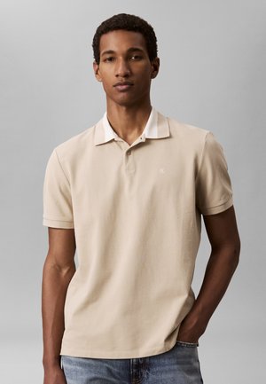 Młody mężczyzna z krótkimi kręconymi włosami, ubrany w beżową koszulkę polo i niebieskie dżinsy, stoi z jedną ręką w kieszeni na szarym tle.