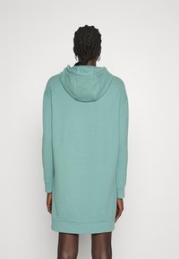 Kvinna med flätat hår klädd i en lös, teal-färgad huvtröjklänning, stående med ryggen mot en enkel vit bakgrund.