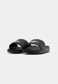 Sandales noires à glissière avec une large sangle arborant un logo de crocodile blanc, fabriquées en matériau lisse avec une semelle robuste.