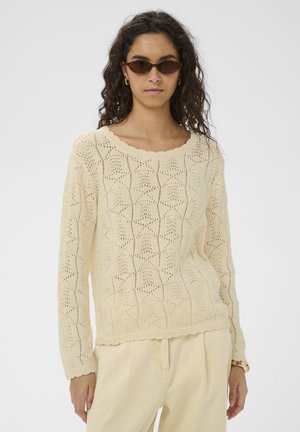 Femme aux cheveux bouclés portant des lunettes de soleil marron, un pull en maille texturée crème, un pantalon crème et des boucles d'oreilles créoles dorées, debout dans une posture neutre.