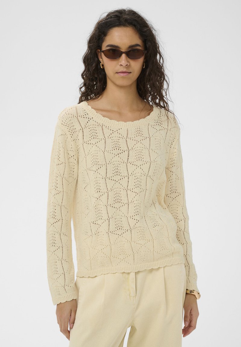 Femme aux cheveux bouclés portant des lunettes de soleil marron, un pull en maille texturée crème, un pantalon crème et des boucles d'oreilles créoles dorées, debout dans une posture neutre.