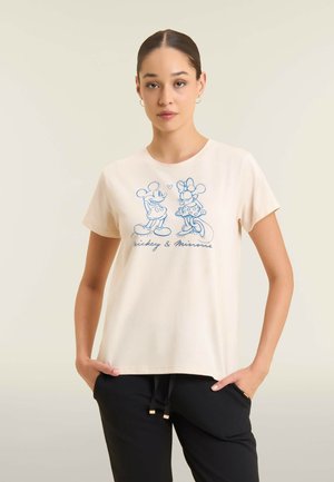 Mujer con cabello oscuro que lleva una camiseta color crema con dibujos en línea azul de Mickey y Minnie Mouse, combinada con pantalones negros, de pie con las manos en los bolsillos.