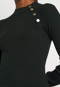 Haut long à manches longues côtelé vert foncé avec un col rond et quatre boutons dorés décoratifs le long de la couture de l'épaule gauche.