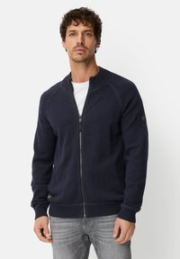 Navy zip-up knit cardigan with a high collar, ribbed cuffs and waistband, and a subtle logo accent on the sleeve. Soft texture.