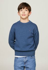 Un maglione blu a maglia con scollatura rotonda e polsini a costine, caratterizzato da un piccolo logo sul petto. Indossato con jeans in denim blu chiaro.