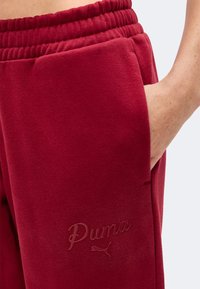 Gros plan sur un pantalon de survêtement rouge Puma avec une ceinture élastique et une main reposant dans la poche latérale.