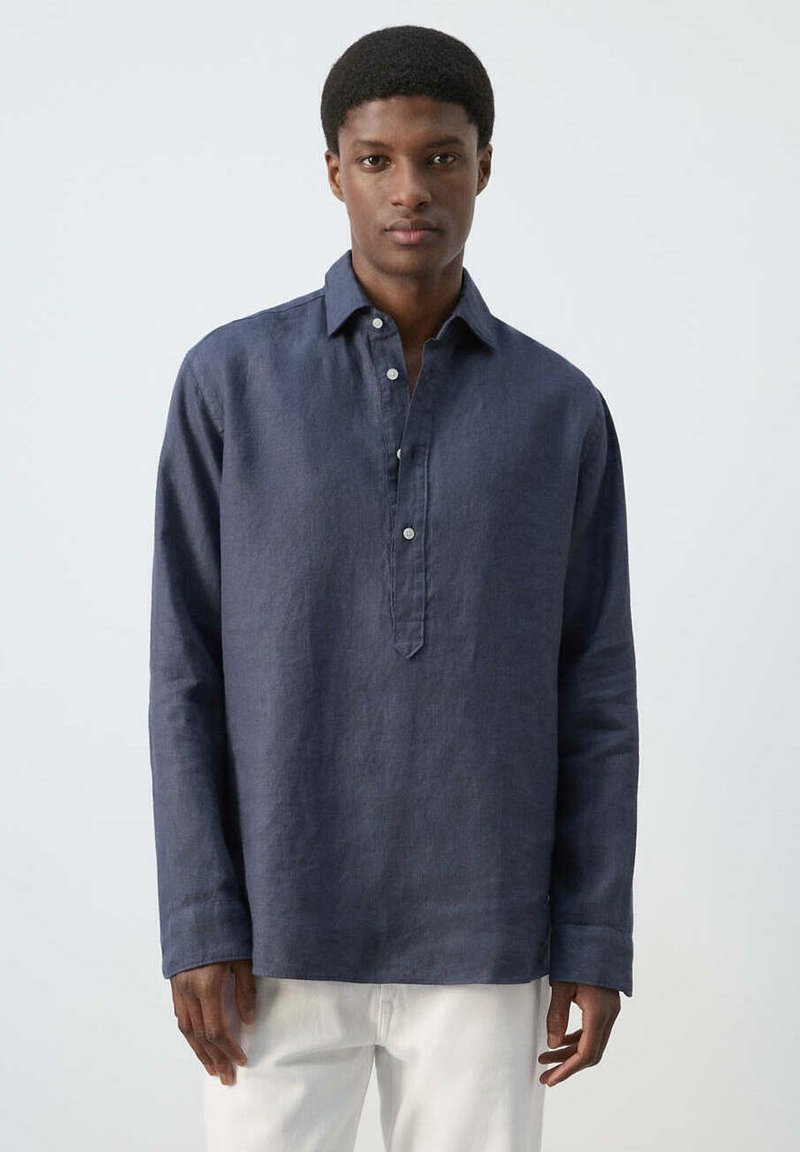 Homme portant une chemise en lin bleu foncé à manches longues avec patte de boutonnage partielle et pantalon blanc, debout devant un fond clair.