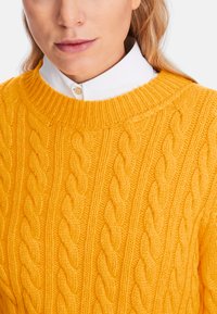 Gelber gestrickter Pullover mit Zopfmuster, ausgestattet mit einem gerippten Rundhalsausschnitt und strukturiertem Garn. Übereinander getragen über einem weißen Hemd mit Kragen.