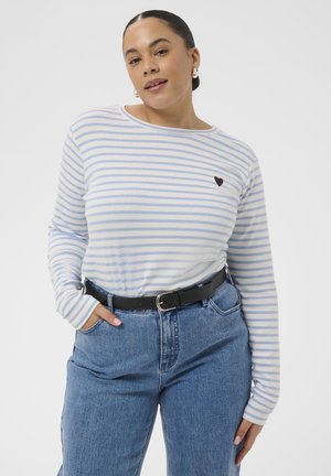 Femme portant une chemise à manches longues rayée bleu clair et blanc avec un petit détail en forme de cœur noir, rentrée dans un jean bleu taille haute avec une ceinture noire.