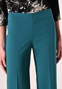 Teal wijde broek met een gladde, matte textuur, voorzien van een aansluitende taille en strakke plooien aan de voorkant, gecombineerd met een top met een bloemenpatroon.