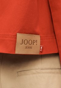 Δερμάτινο λογότυπο με το κείμενο «JOOP! JEANS» στο τελείωμα ενός κόκκινου ρούχου πάνω από μπεζ παντελόνι με εμφανή πίσω τσέπη.