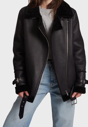 Person trägt eine schwarze Lammfelljacke mit Schnallen an Ärmeln und Saum, hellblaue Jeans und ein graues Oberteil, steht vor einem einfarbigen Hintergrund.