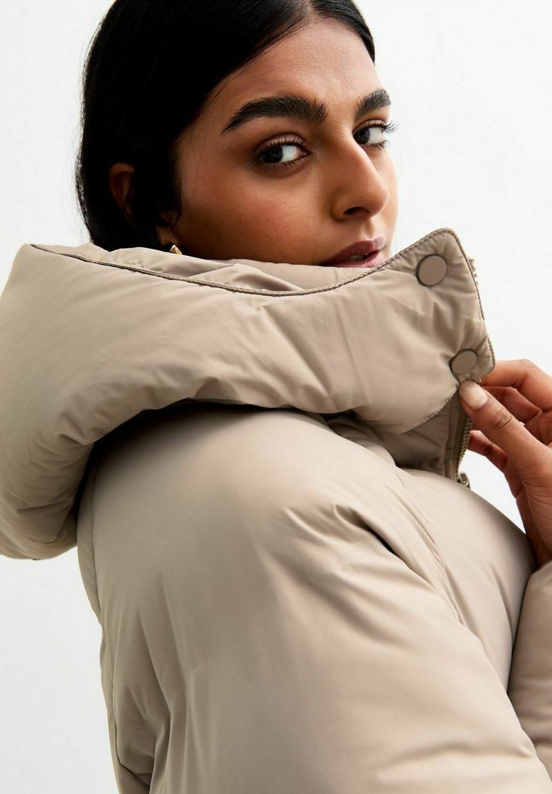 Beżowa kurtka puchowa z wysokim kołnierzem, z zapięciem na napy i gładką teksturą. Modelka prostuje kołnierz, spoglądając w tył.