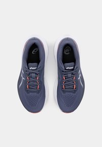 Baskets de sport grises avec une tige en maille, des lacets orange contrastants et une semelle blanche. Présence d'un logo sur la languette et de trous de ventilation bien visibles.