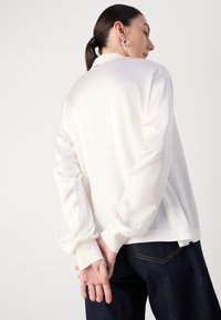 Blouse en satin blanc avec des manches longues et amples et des poignets ajustés. Texture lisse, design minimaliste, avec un col ; associée à un jean en denim foncé.