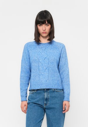 Ung kvinde med mørkt hår iført lyseblå kabelstrikket sweater og blå jeans, stående op ad en ensfarvet hvid baggrund, med ansigtet fremad.