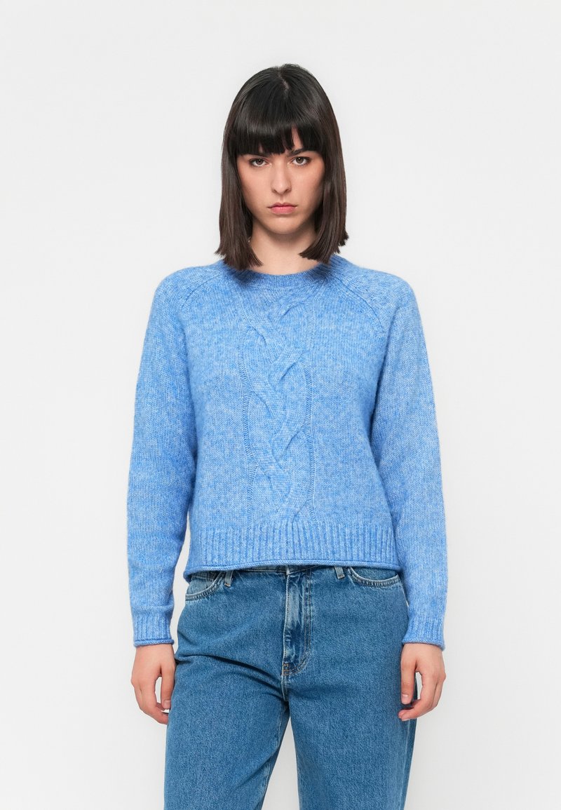 Junge Frau mit dunklem Haar trägt einen hellblauen Zopfmuster-Pullover und blaue Jeans, steht vor einem schlichten weißen Hintergrund und schaut nach vorne.