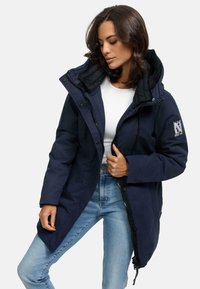 Veste longue bleu marine avec fermeture éclair et patte de boutonnage, capuche et texture matelassée. Comprend un détail en patch sur le bras gauche.
