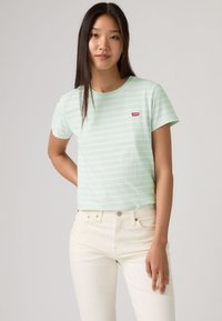 Νεαρή γυναίκα με μακριά σκούρα μαλλιά, που φοράει μια ανοιχτοπράσινη ριγέ μπλούζα και εκρού τζιν, στέκεται μπροστά σε ένα απλό φόντο.