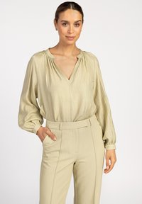 Helle olivgrüne Bluse mit langen Ärmeln, V-Ausschnitt und gerafftem Halsdetail, kombiniert mit passend zugeschnittenen Hosen mit seitlichen Taschen.