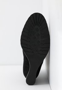 Chaussure en daim noir avec une semelle en caoutchouc nervurée. Présente une indication de taille visible (37) et un design de bout lisse. Positionnée verticalement sur un fond blanc.