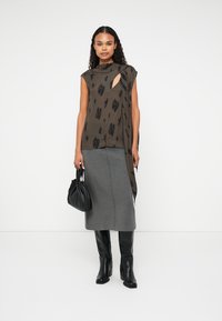 Blouse sans manches kaki avec un motif floral noir, associée à une jupe midi grise et des bottes noires hauteur genou, tenant un sac à main noir.