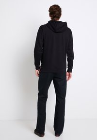 Felpa nera con cappuccio a marsupio, cappuccio con cordino e polsini a coste, abbinata a pantaloni scuri e scarpe nere, vista da dietro.