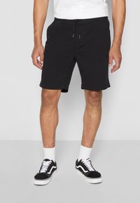 Homme portant un short noir avec cordon de serrage, des chaussettes blanches de type crew et des baskets noires basses avec des lacets blancs, se tenant sur un sol uni.