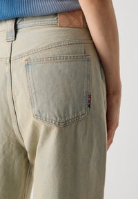Jean en denim délavé avec taille haute, présentant une insigne en cuir à l'arrière et des accents brodés rouge et bleu près de la poche.