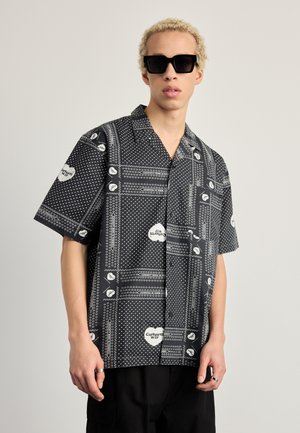 Camisa negra de mangas cortas con un diseño estampado de lunares blancos y motivos de corazones. El material parece ligero y suave.