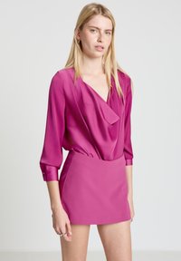 Fuchsiabluse mit drapiertem Frontteil und V-Ausschnitt, kombiniert mit einem passenden fuchsiafarbenen Minirock. Glatte Textur, lange Ärmel mit Knopfdetails.