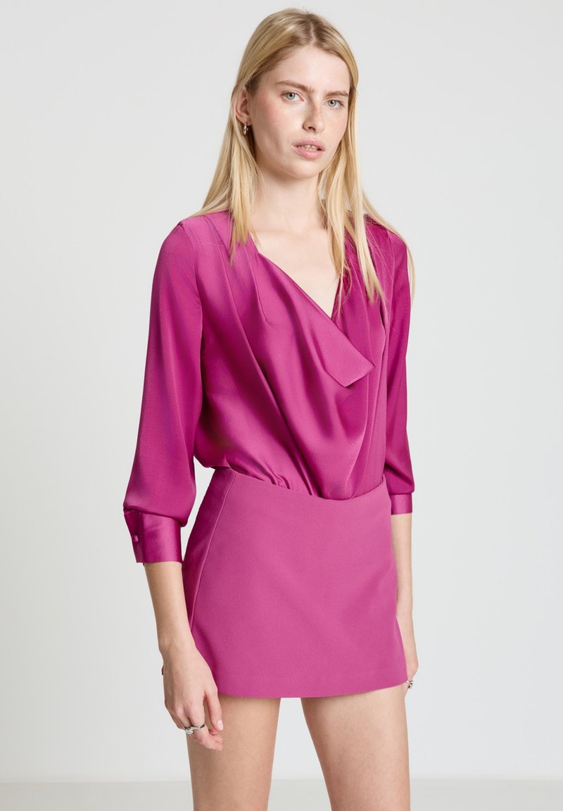 Fuchsiabluse mit drapiertem Frontteil und V-Ausschnitt, kombiniert mit einem passenden fuchsiafarbenen Minirock. Glatte Textur, lange Ärmel mit Knopfdetails.